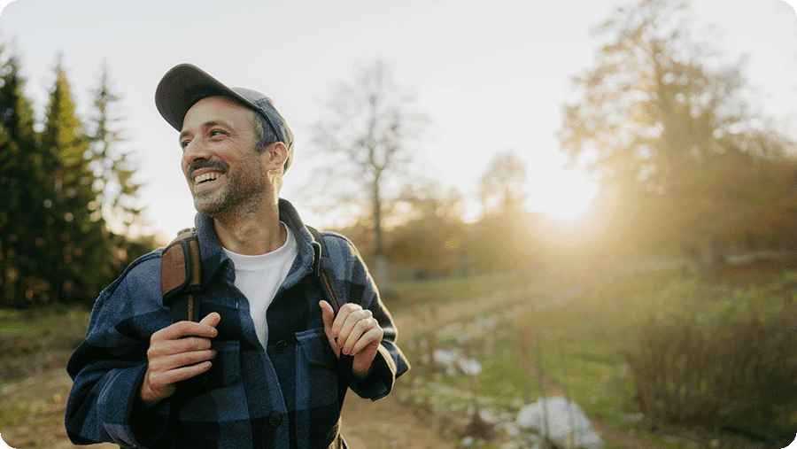 Smiling man wearing a plaid jacket and baseball cap hiking outdoors with a backpack, walking along a sunlit trail surrounded by trees in early autumn.